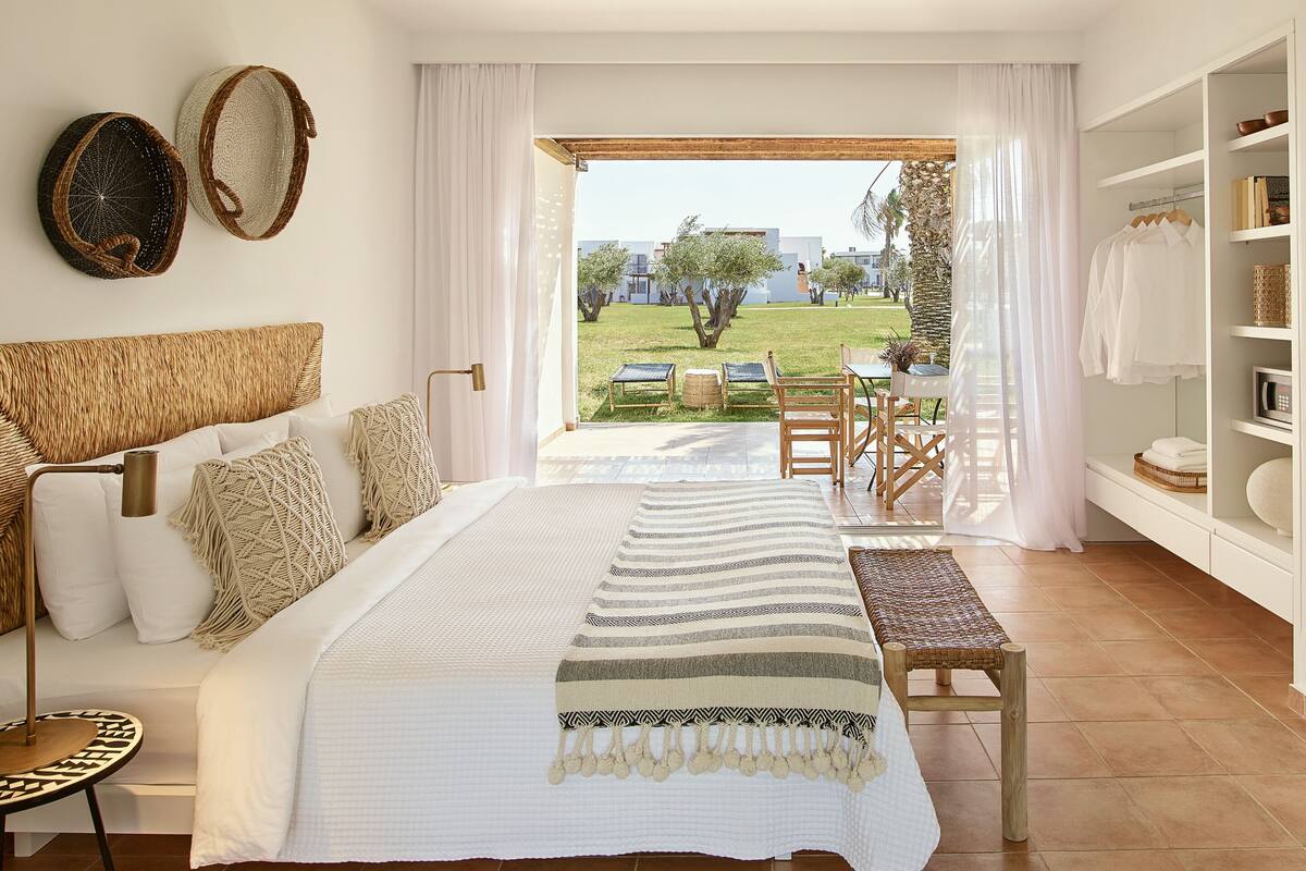 A spacious bedroom with a large bed, a bedside table, a bench, and a view of a garden through a large window.