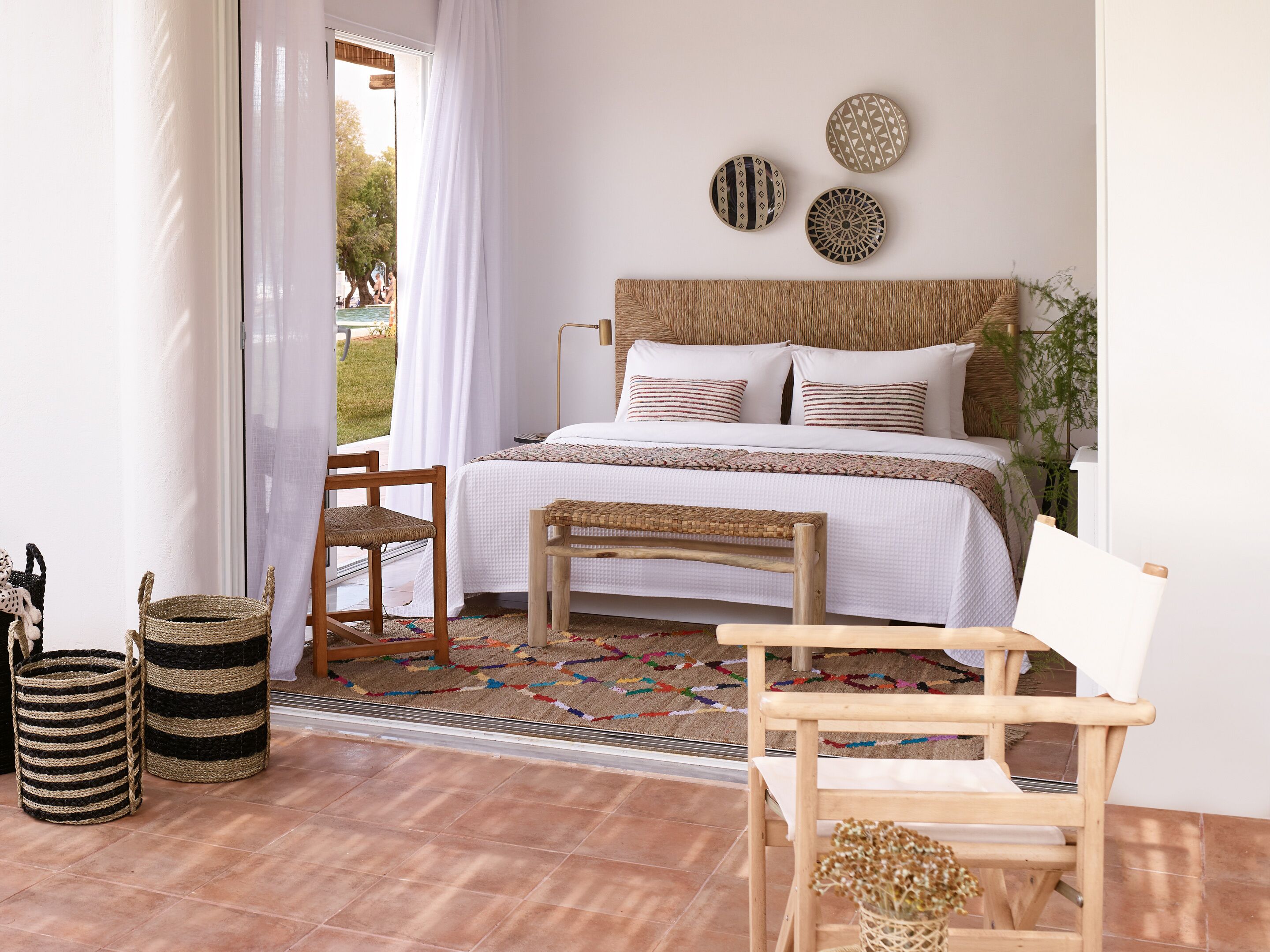 A bedroom with a bed, two wicker chairs, a small table, and a view of the outdoors.