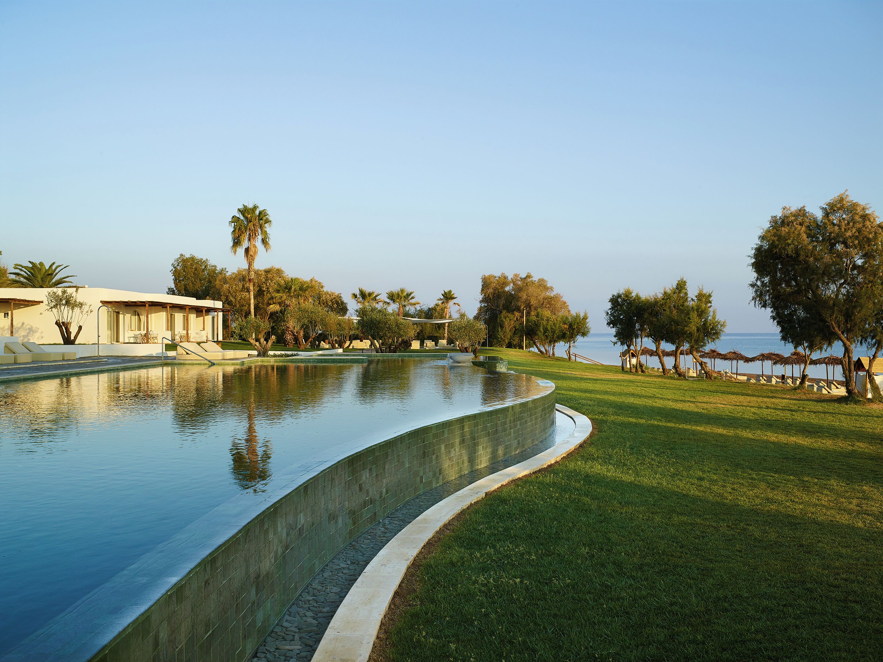 A swimming pool with a curved edge, surrounded by well-maintained grass and trees, with a view of the sea in the background.