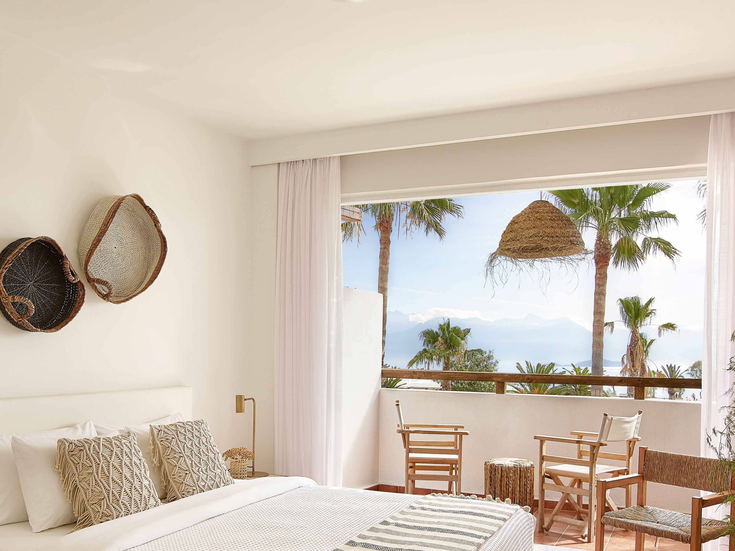 Una camera da letto con un letto, due cuscini in macramè, un comodino, una lampada e vista su palme e montagne attraverso una finestra con balcone.