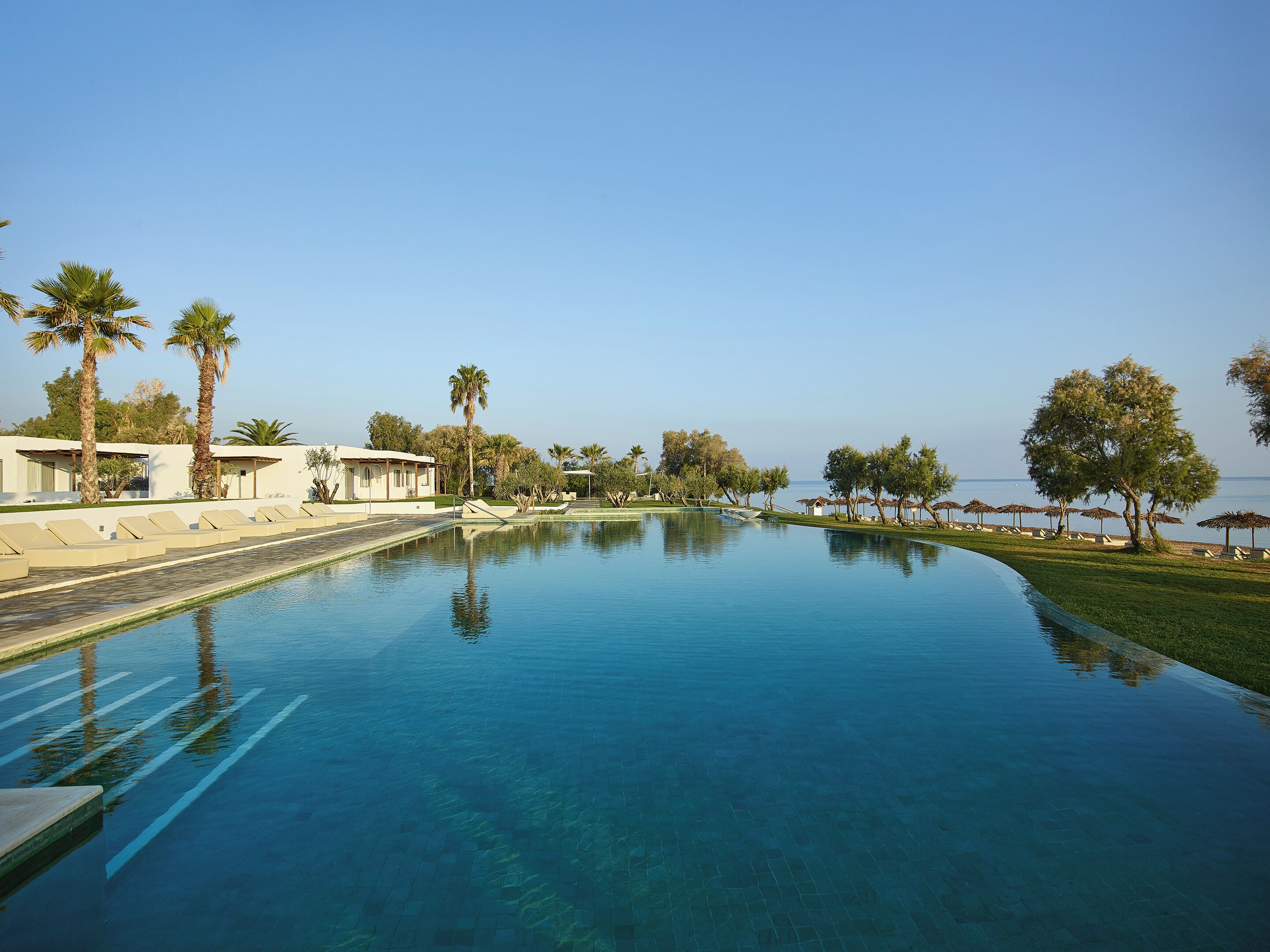 A swimming pool with lounge chairs and palm trees, surrounded by a clear sky and a view of the sea.