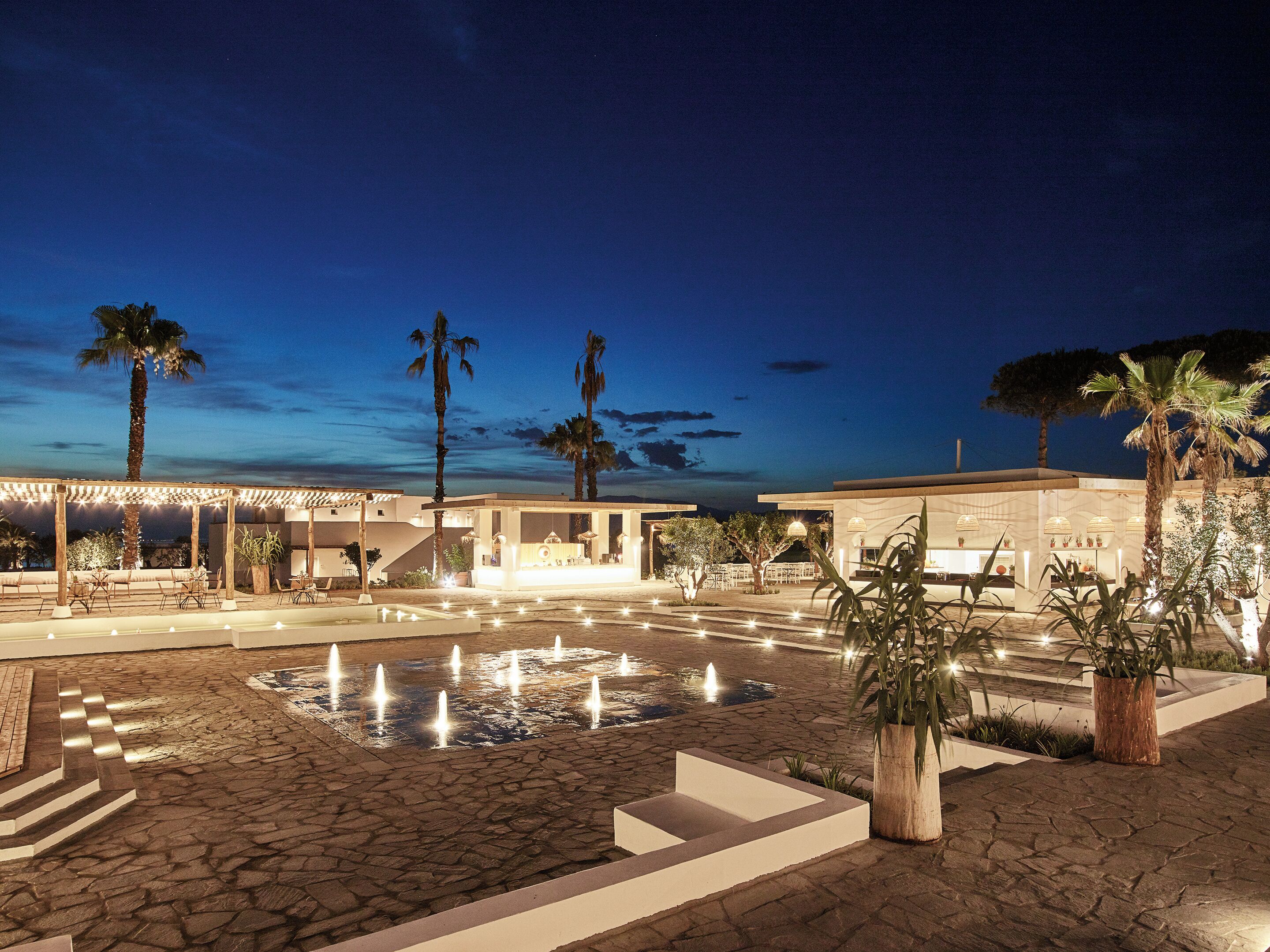 A nighttime outdoor area with a water feature, palm trees, and a covered seating area.