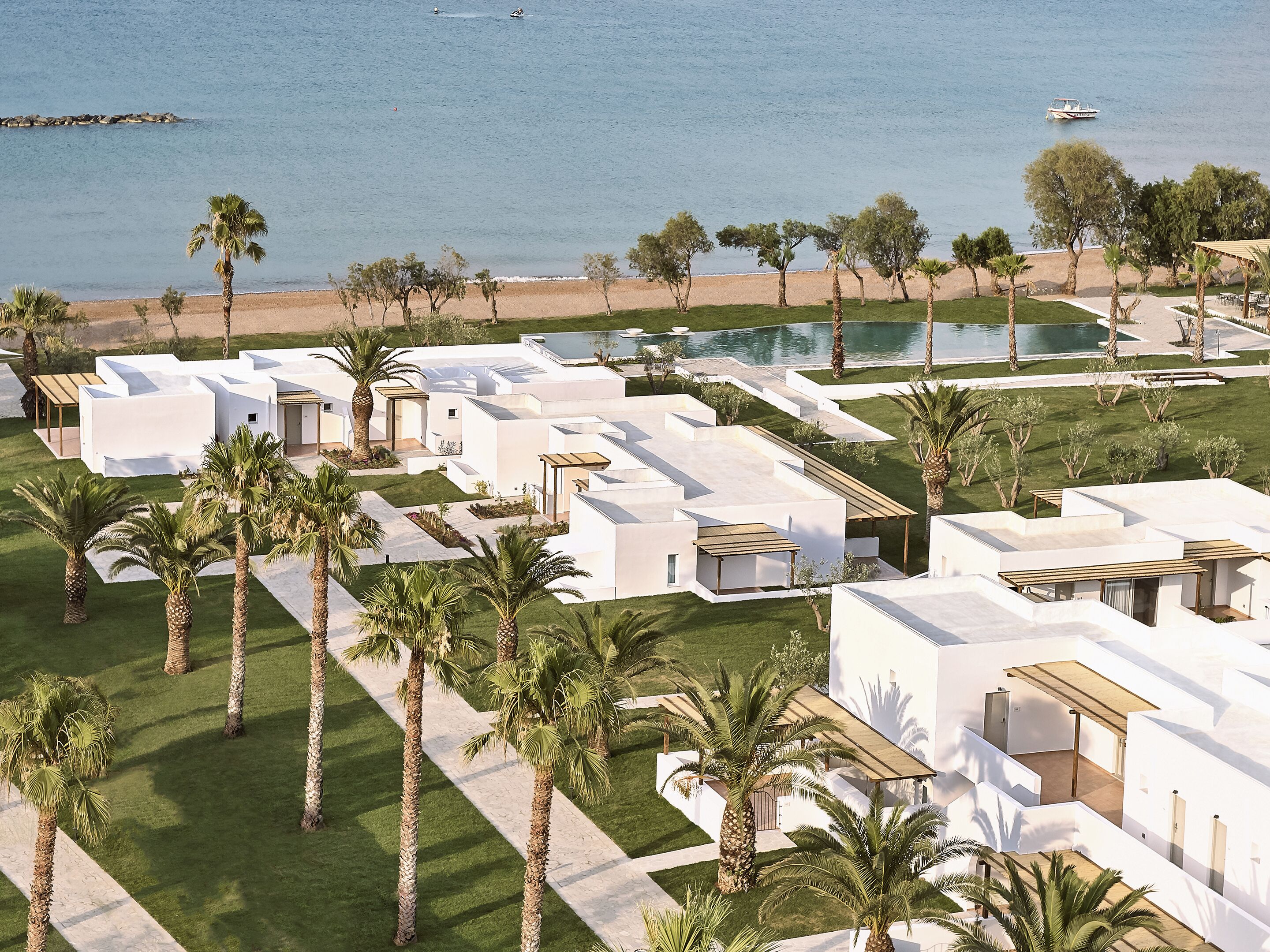 A modern resort with white buildings, palm trees, a beach, and a pool.