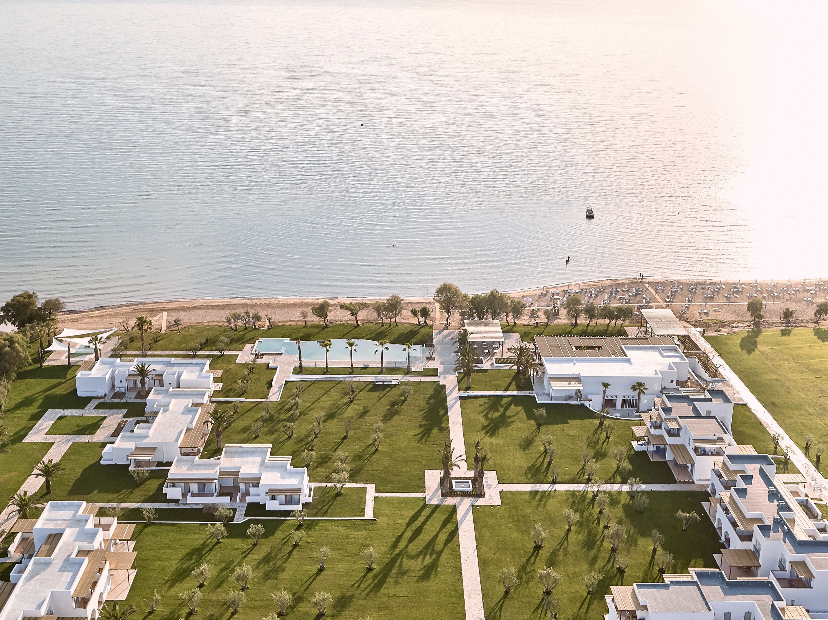 An aerial view of a resort with multiple white buildings, a central pool area, and a beach in the background.