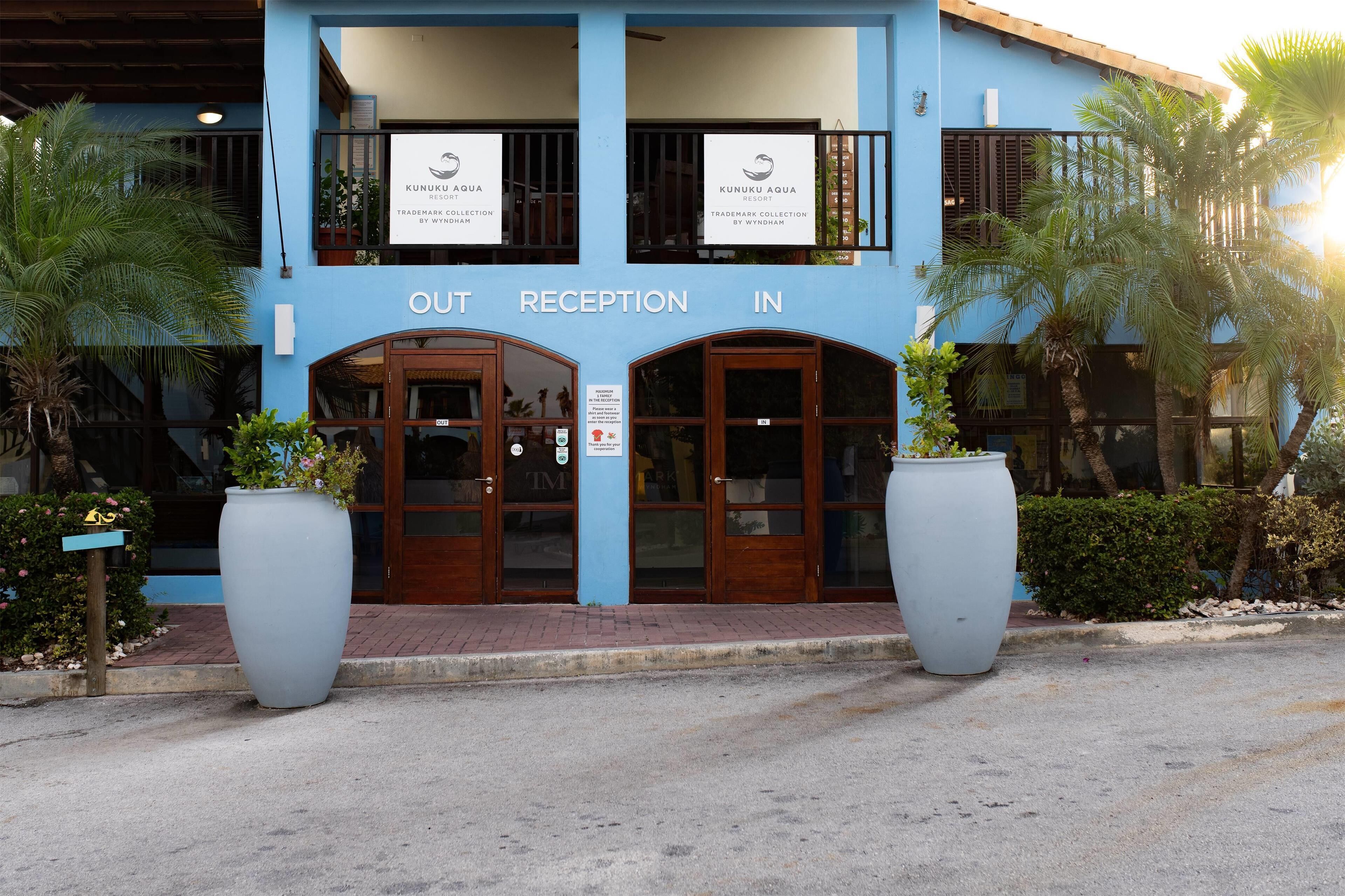 Ein Gebäude mit blauer Fassade, zwei große Kübelpflanzen und Schilder mit den Aufschriften 'AUS RECEPTION IN' und 'IN RECEPTION'.