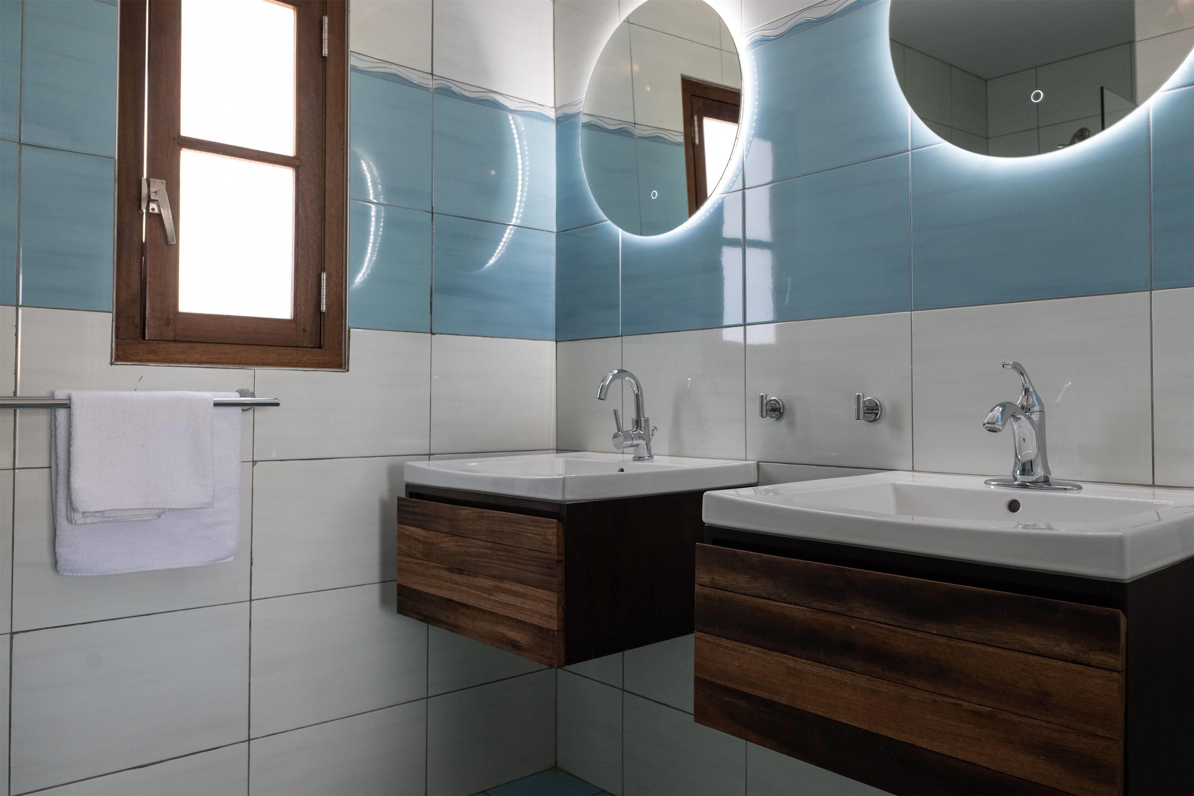 A modern bathroom with two sinks, wooden cabinets, and a large mirror with circular lighting.