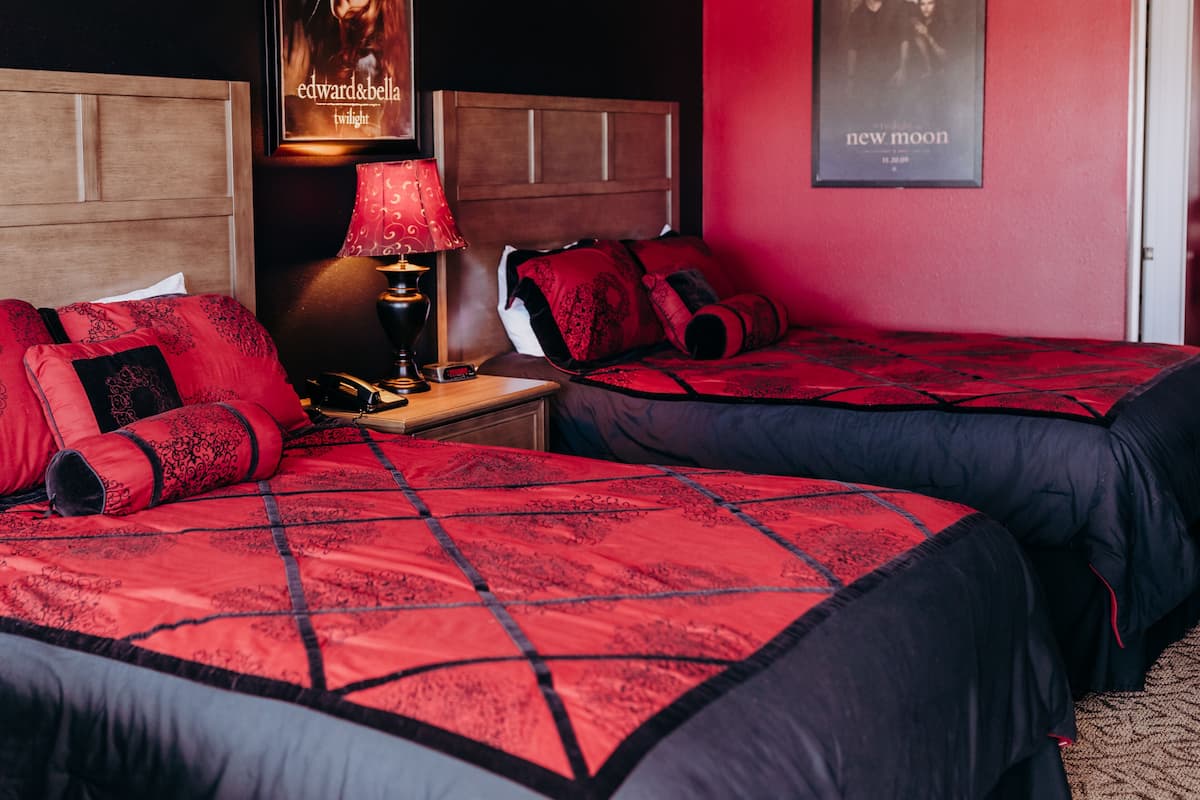 Quarto de hotel com duas camas, uma mesa de cabeceira, um abajur e cartazes nas paredes.