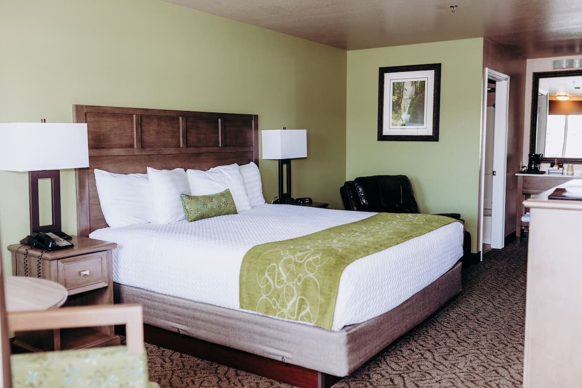 Quarto de hotel com uma cama grande, duas luminárias de cabeceira, uma cadeira, uma escrivaninha e um espelho.