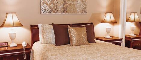 A neatly made bed with a patterned bedspread, two bedside tables with lamps, a wall clock, and a decorative wall piece.