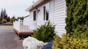 Una casa con fachada blanca, un porche y diversas plantas, incluyendo flores blancas, flores azules y arbustos verdes.