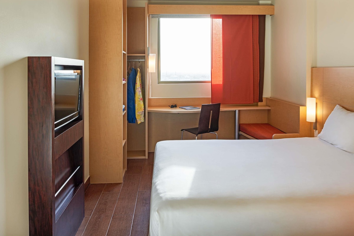 A hotel room with a bed, a desk with a chair, a TV, and a wardrobe.