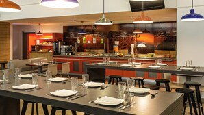 Restaurante com várias mesas postas para os clientes, iluminação pendente no teto e uma área de cozinha visível ao fundo.