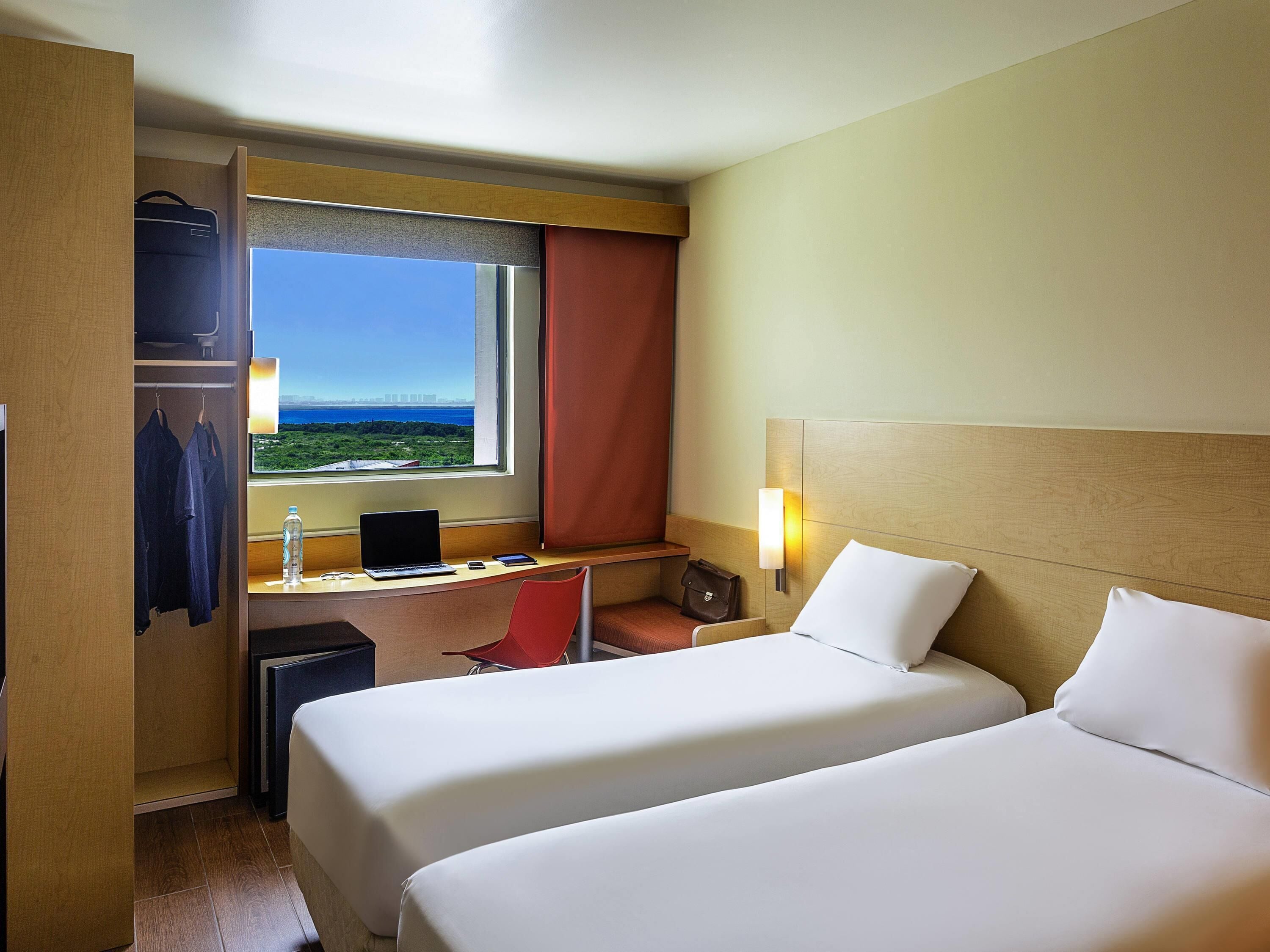 Habitación de hotel con dos camas, un escritorio con una computadora portátil, una silla, un armario y una ventana con vistas.