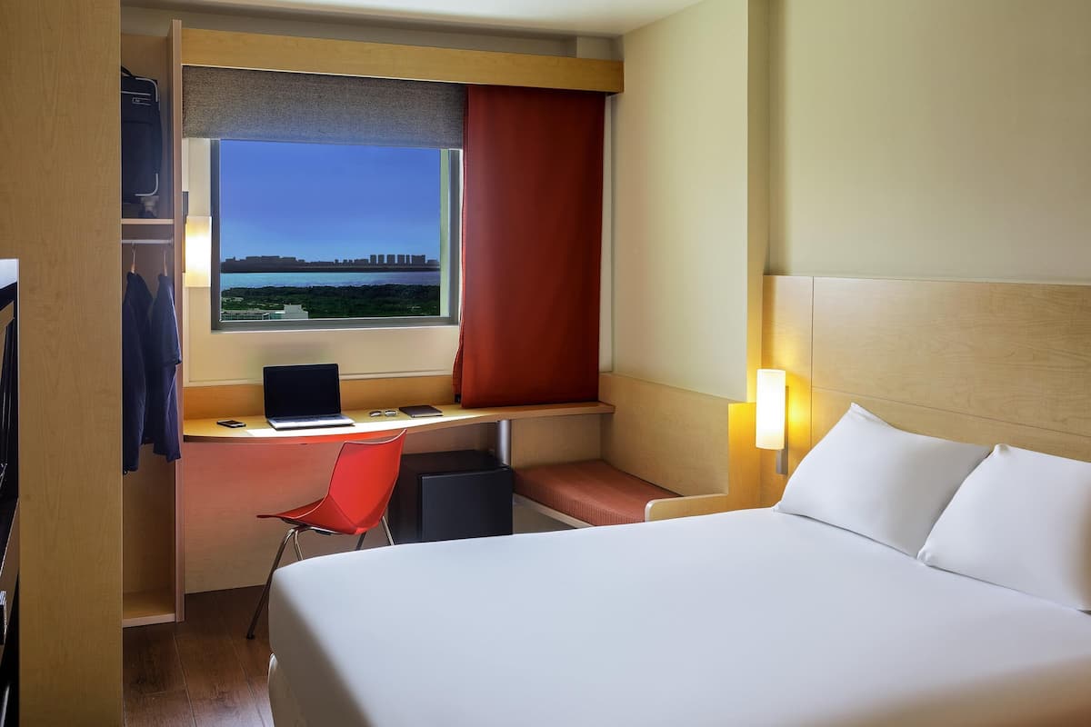 Habitación de hotel con cama, escritorio con una computadora portátil, silla, ventana con vista a un paisaje urbano y armario.