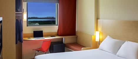 Habitación de hotel con cama, escritorio con una computadora portátil, silla, ventana con vista a un paisaje urbano y armario.