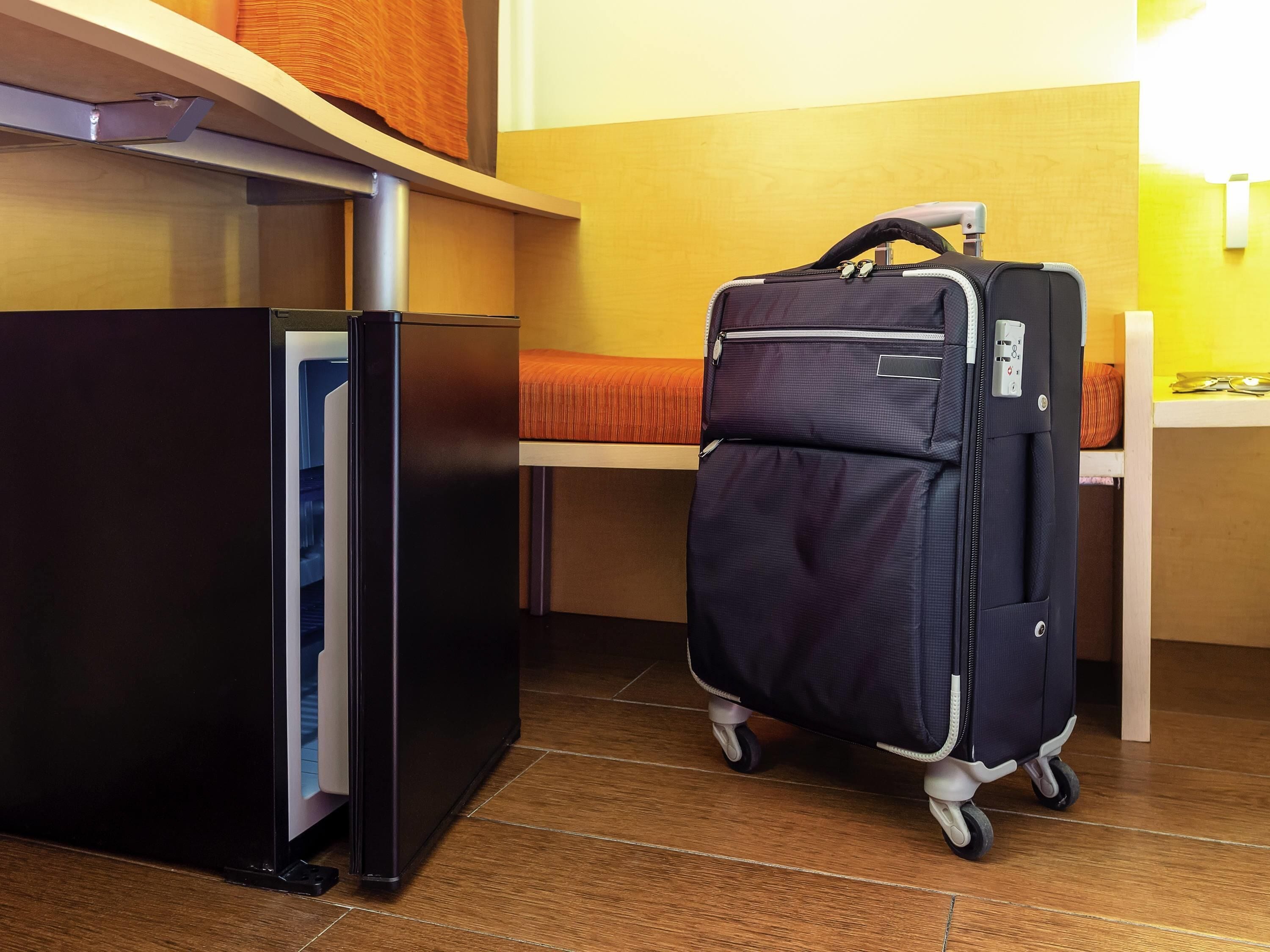 A compact room with a black refrigerator, a suitcase on wheels, and a bed with a striped blanket.