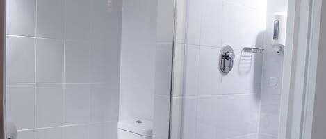 A modern bathroom with white tiles, a toilet, a glass-enclosed shower, and a small trash bin.