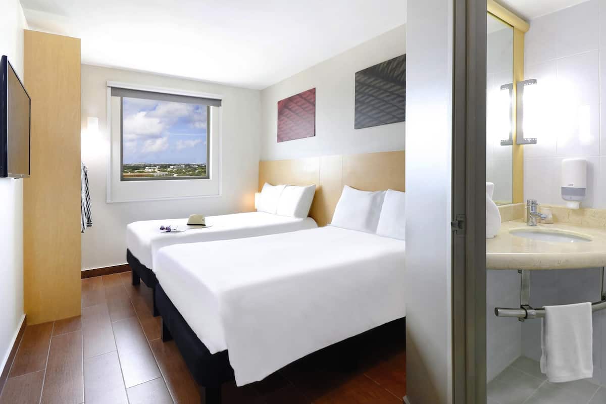 Habitación de hotel con dos camas, un ventanal con vistas a la ciudad, baño con lavamanos y espejo, y un perchero para toallas.
