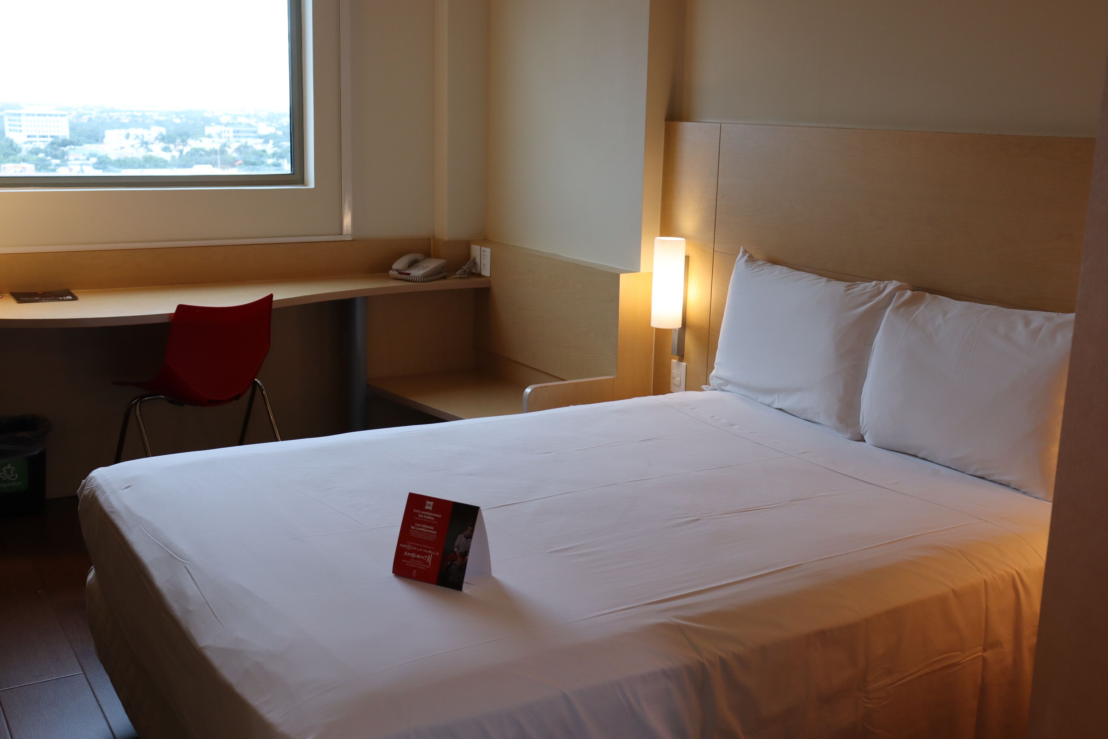Une chambre d’hôtel équipée d’un lit, d’un bureau, d’une chaise, d’une lampe et d’une fenêtre donnant sur la ville.