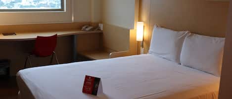 Una habitación de hotel con cama, escritorio, silla, lámpara y ventana con vista a la ciudad.