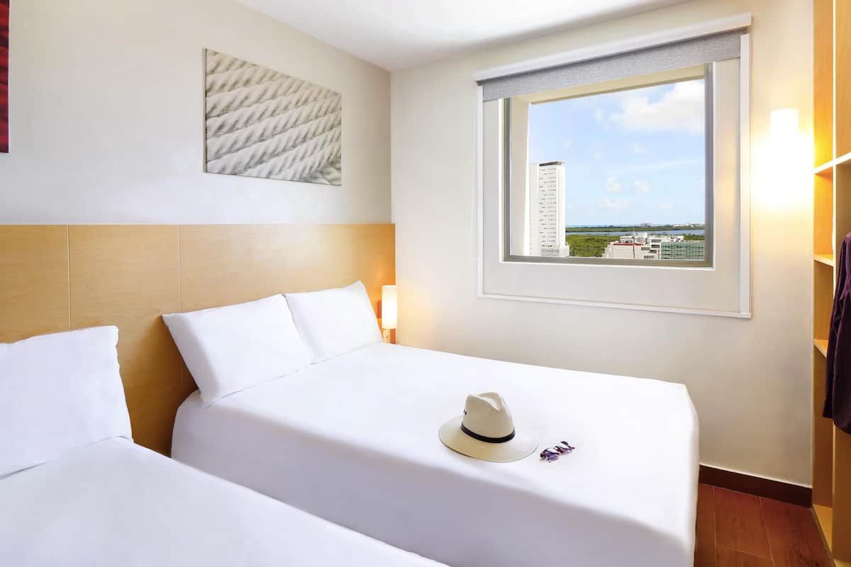 Habitación de hotel con dos camas, una ventana con vista a la ciudad, una obra de arte enmarcada en la pared y un cabecero de madera.