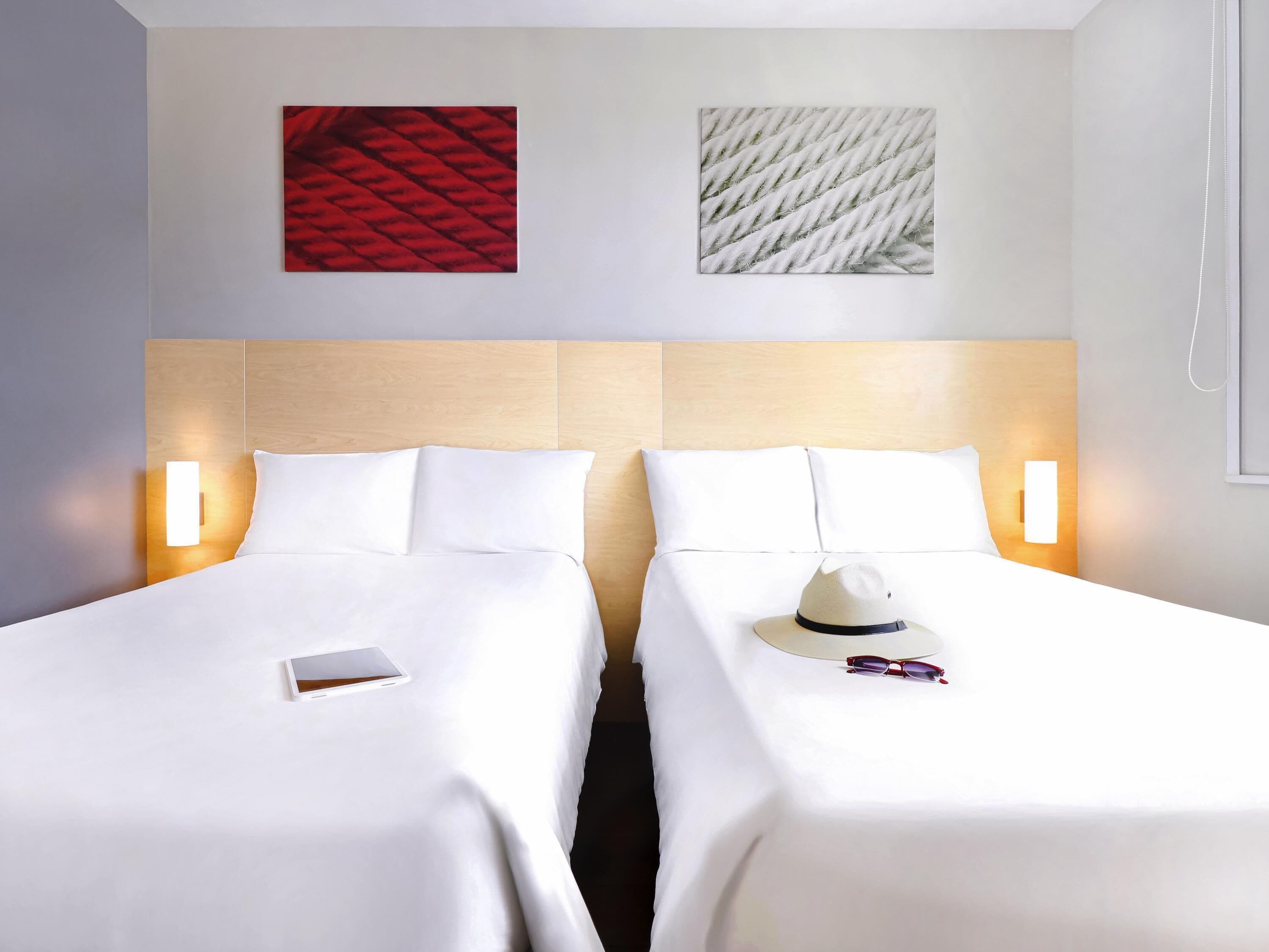 A hotel room with two beds, each with white linens and pillows. There are wall-mounted light fixtures on either side of the beds.