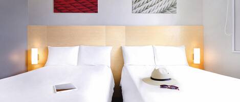 A hotel room with two beds, each with white linens and pillows. There are wall-mounted light fixtures on either side of the beds.
