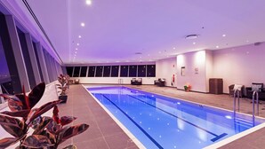Una amplia piscina interior con suelo de baldosas, rodeada de ventanas y plantas de interior.