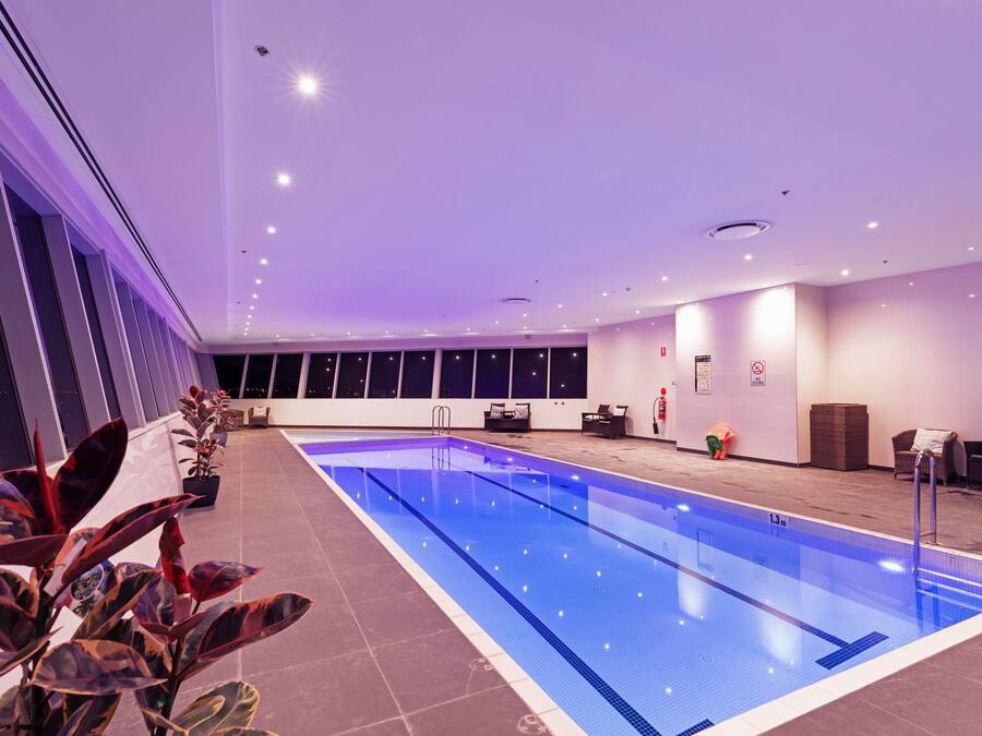 A large indoor swimming pool with a tiled floor, surrounded by windows and indoor plants.