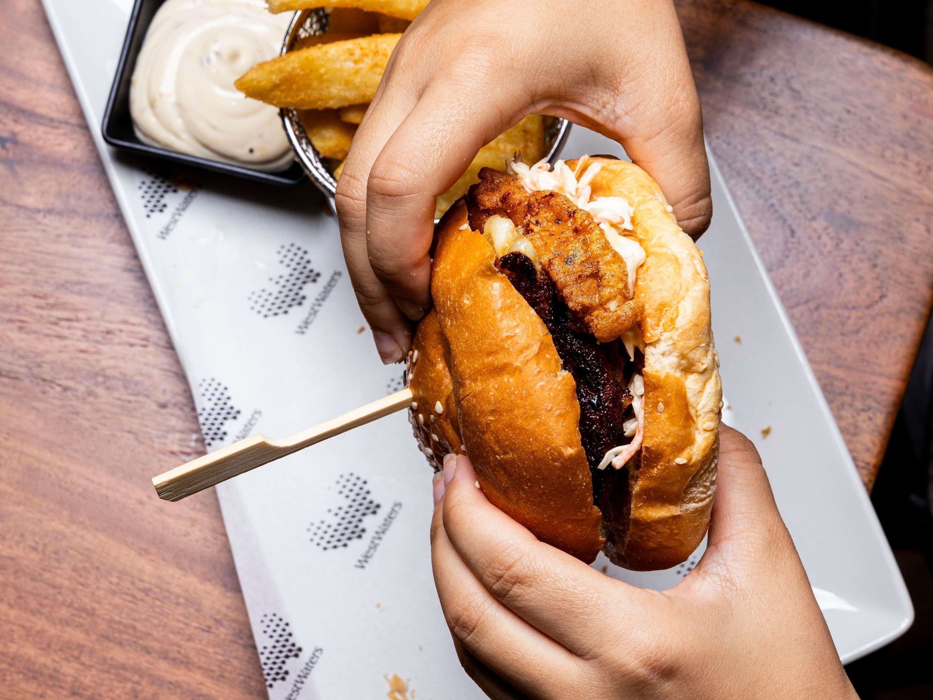 A person holding a sandwich with a toothpick, accompanied by a plate of fries and dipping sauce.