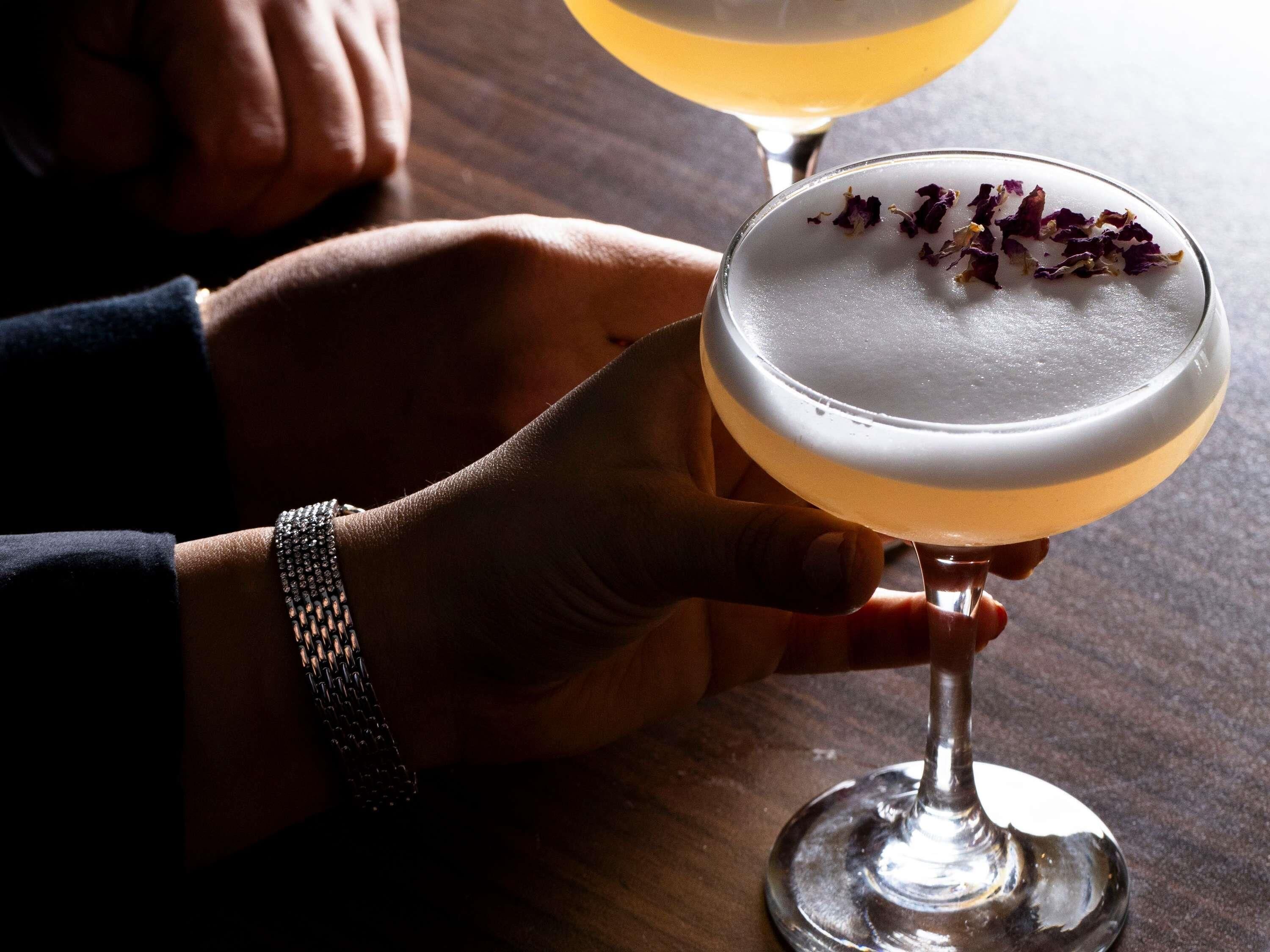 A person holding a cocktail glass with a garnish, and another glass in the background.