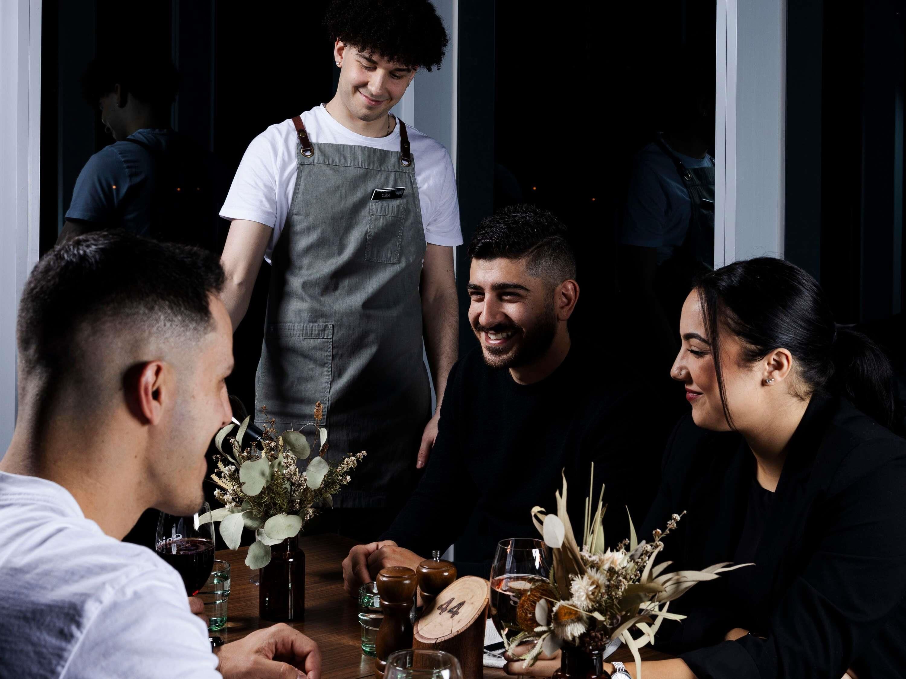 A group of people seated around a table with drinks and flowers, in a dimly lit indoor setting.