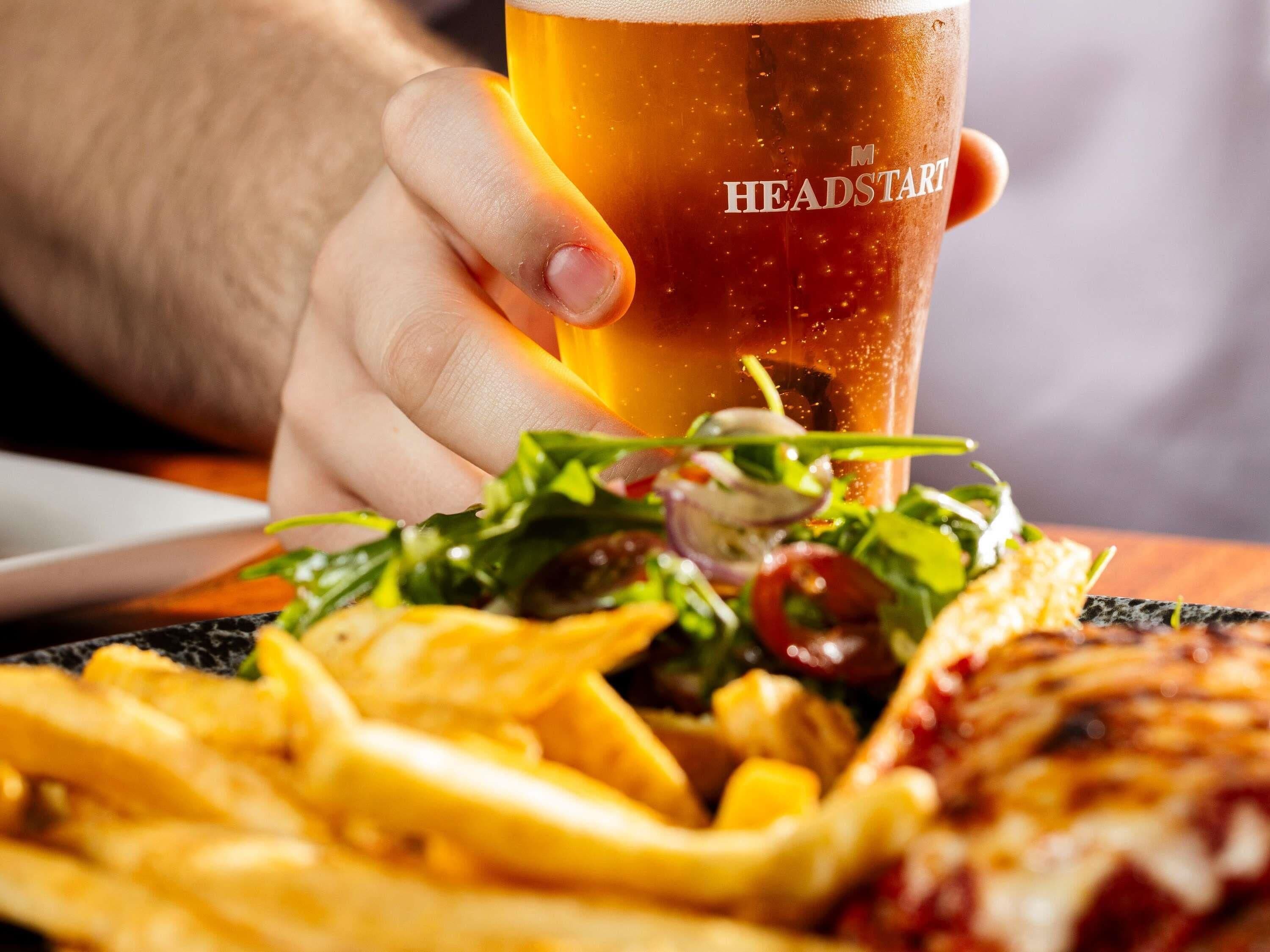 Una persona che tiene un bicchiere di birra con la scritta 'HEADSTART', un piatto di cibo che include patatine fritte e pizza, e un'insalata con verdure a foglia verde e pomodori.