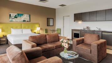 A hotel room with a bed, two sofas, a small table with magazines, a vase with flowers, a painting on the wall, and a kitchenette.
