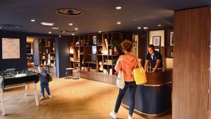 A lobby with a reception desk, wooden shelving with various items, and a foosball table. - ibis Styles Nantes Rezé Aéroport (Reze)