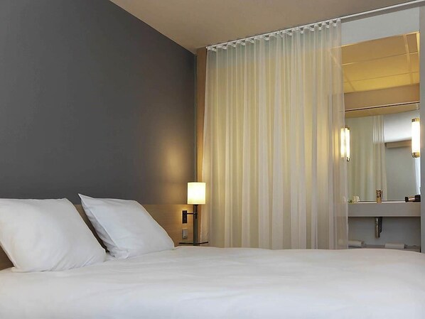 Une chambre d’hôtel avec un lit, une table de chevet, une lampe, un miroir et une fenêtre avec des rideaux légers.