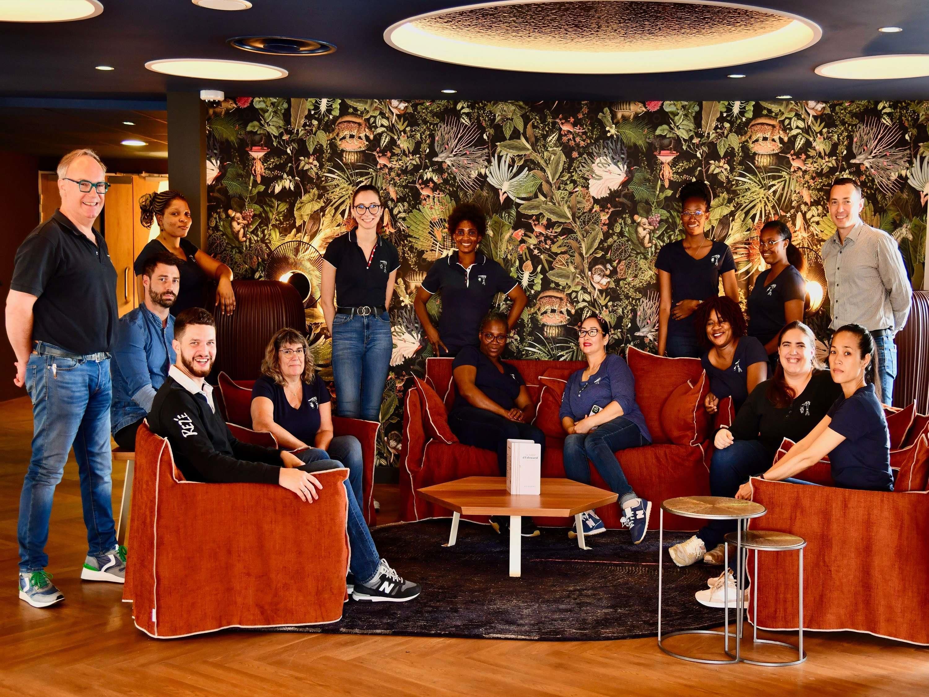 Un groupe de personnes se trouve dans un salon moderne, avec un mur décoré sur le thème des tropiques, des sièges confortables et une petite table basse.
