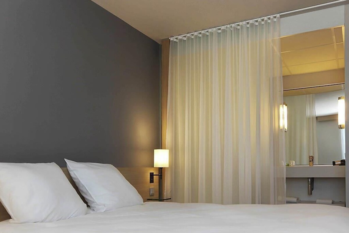 Une chambre d’hôtel avec un lit, une table de chevet avec une lampe, une salle de bain avec un miroir et des rideaux transparents.