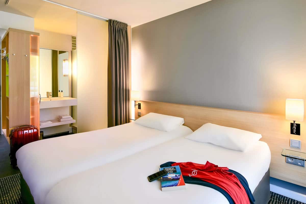 Una habitación de hotel con cama, mesita de noche con lámpara, espejo, armario y maleta.