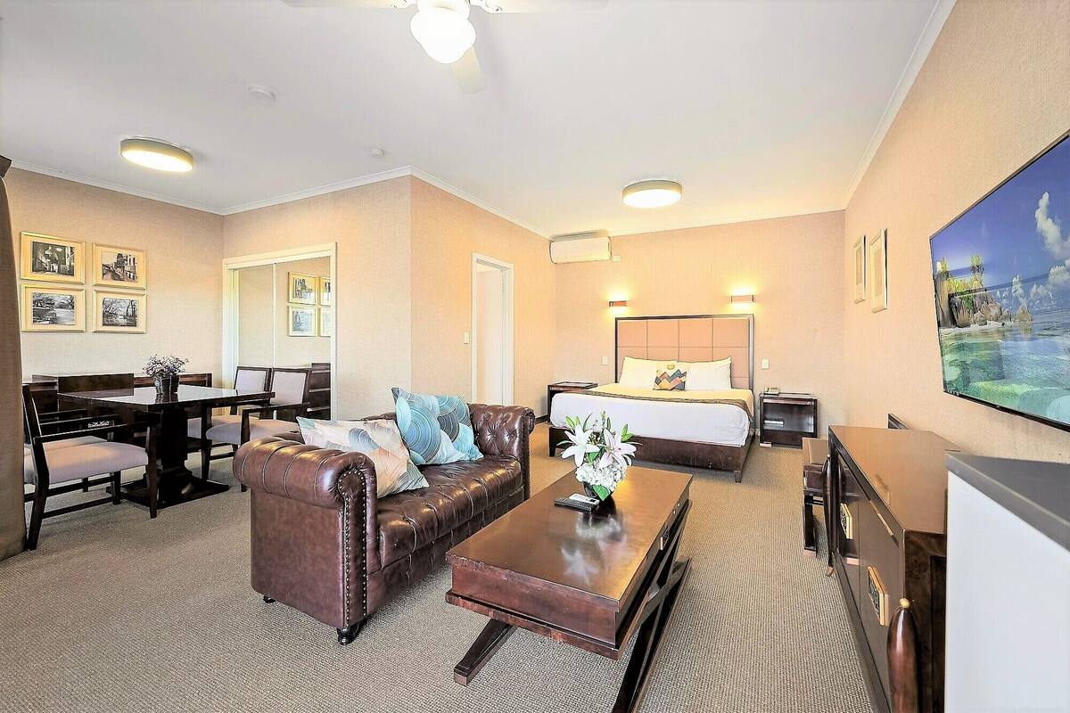 A hotel room with a dining area, a living room with a leather sofa, a bed with a headboard, and a TV.