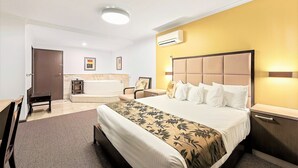 A hotel room with a bed, bedside tables, a desk, a chair, a small table, a bathtub, and an air conditioning unit. - Eltham Gateway Hotel (Eltham)