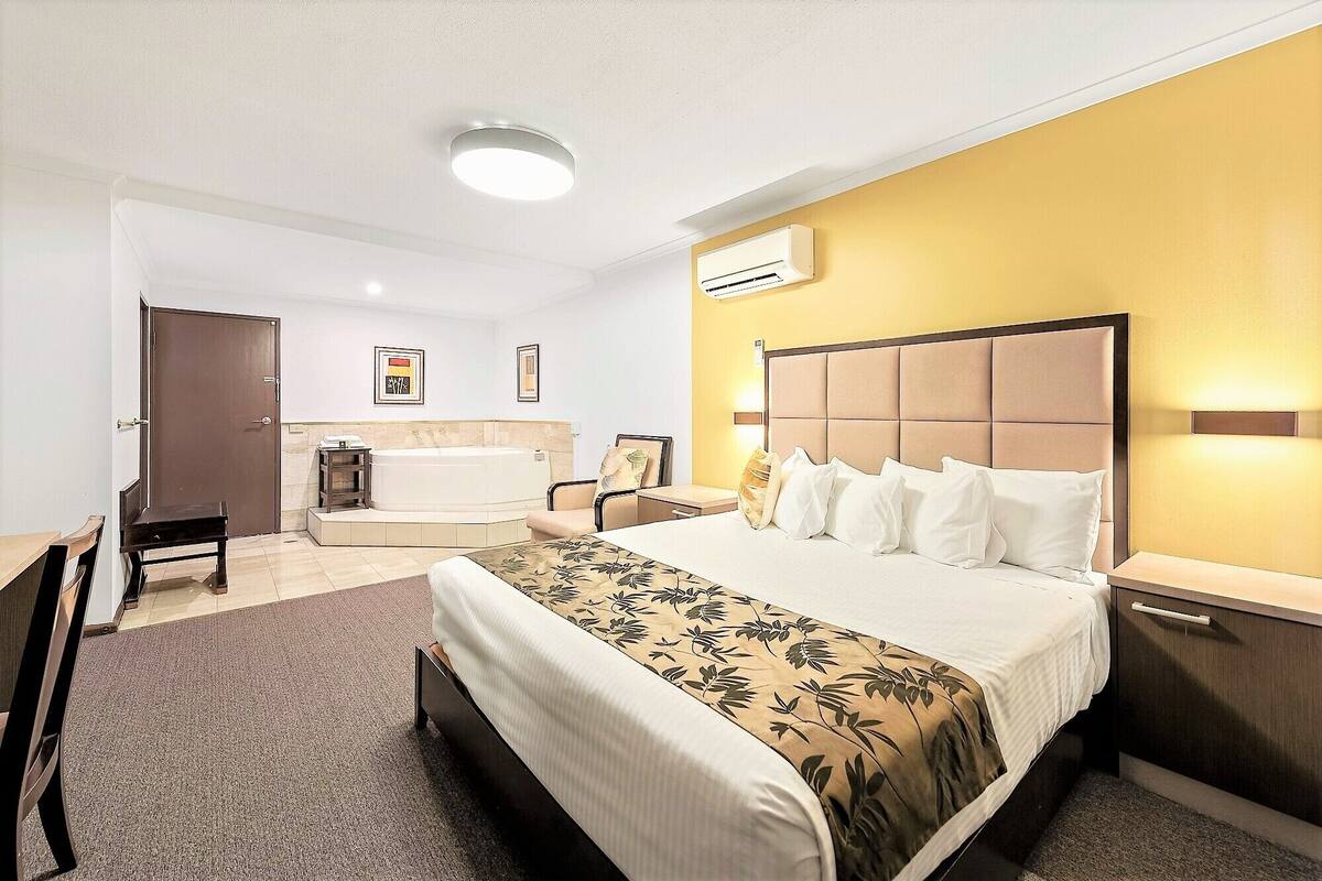 A hotel room with a bed, bedside tables, a desk, a chair, a small table, a bathtub, and an air conditioning unit.