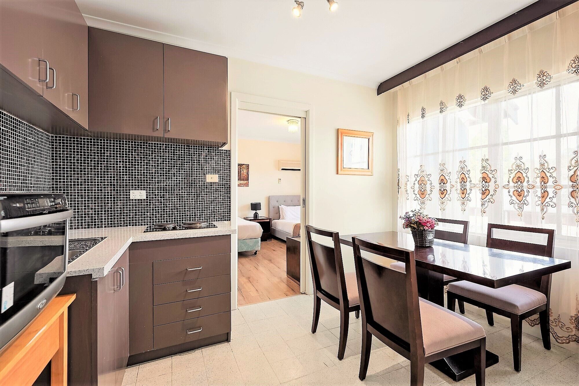 Una cocina moderna con armarios oscuros, salpicadero de azulejos y un comedor con muebles de madera y un amplio ventanal con cortinas decorativas.