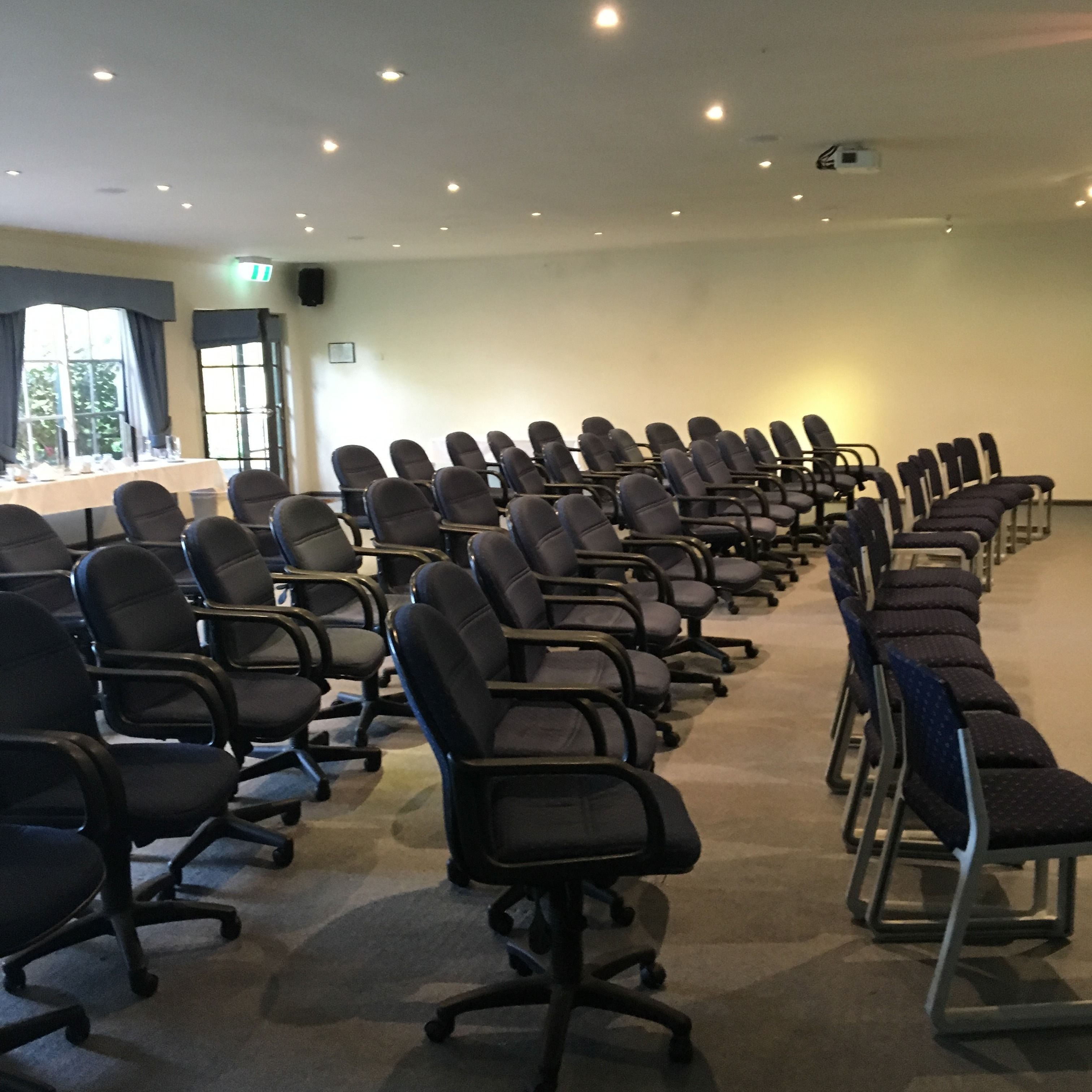 Une salle de conférence avec des rangées de chaises de bureau noires, une longue table recouverte d’une nappe blanche et un écran de projection à l’avant.