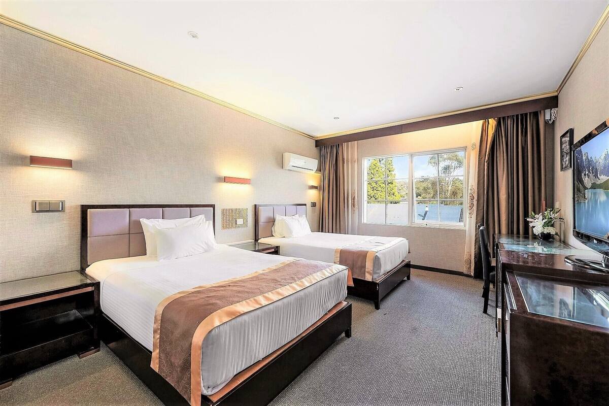 A hotel room with two beds, a TV, and a window with a view of trees.