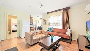 A living room with a sofa, a coffee table, and a television. There is a kitchen area visible in the background. - Eltham Gateway Hotel (Eltham)