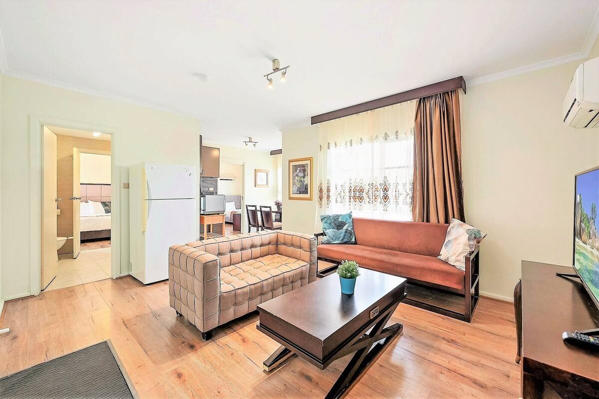 A living room with a sofa, a coffee table, and a television. There is a kitchen area visible in the background.
