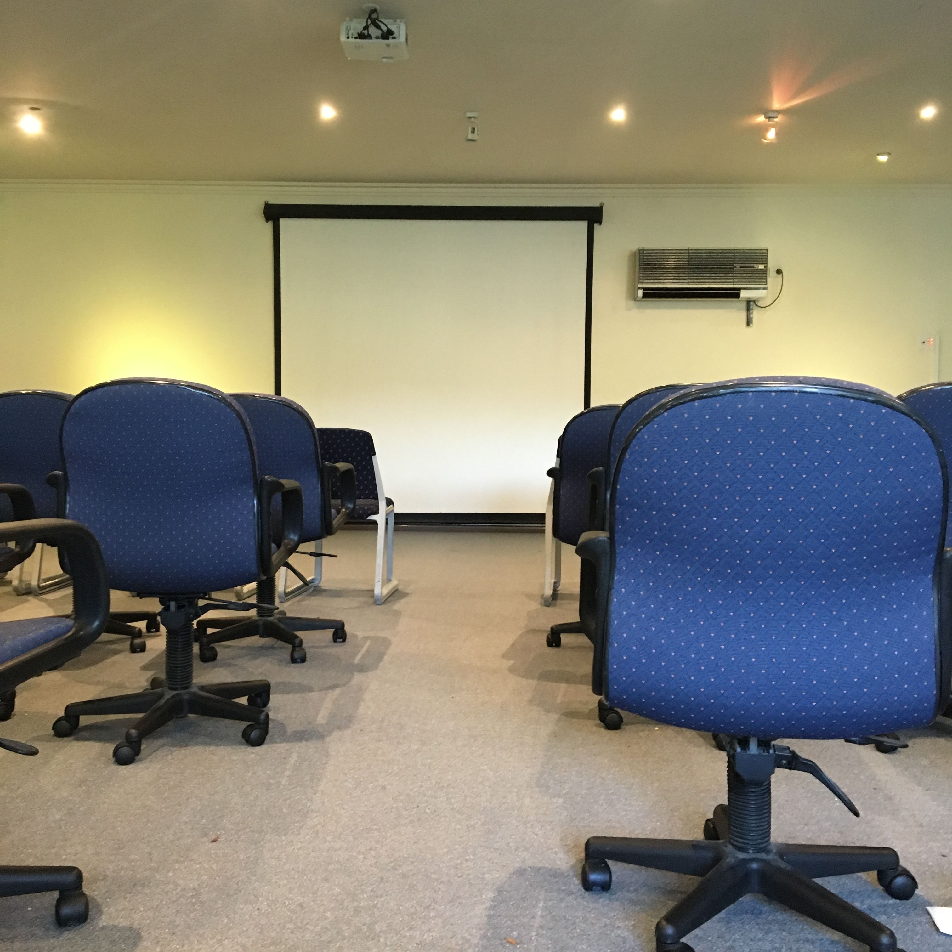 Una sala de conferencias con filas de sillas de oficina azules, una pantalla de proyector y un equipo de aire acondicionado en la pared.