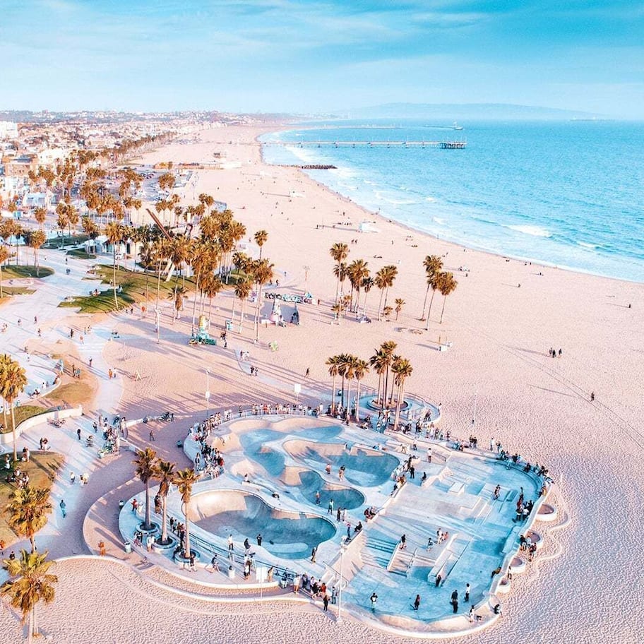 An aerial view of a beach with a large pool area, palm trees, and a coastal cityscape in the background.