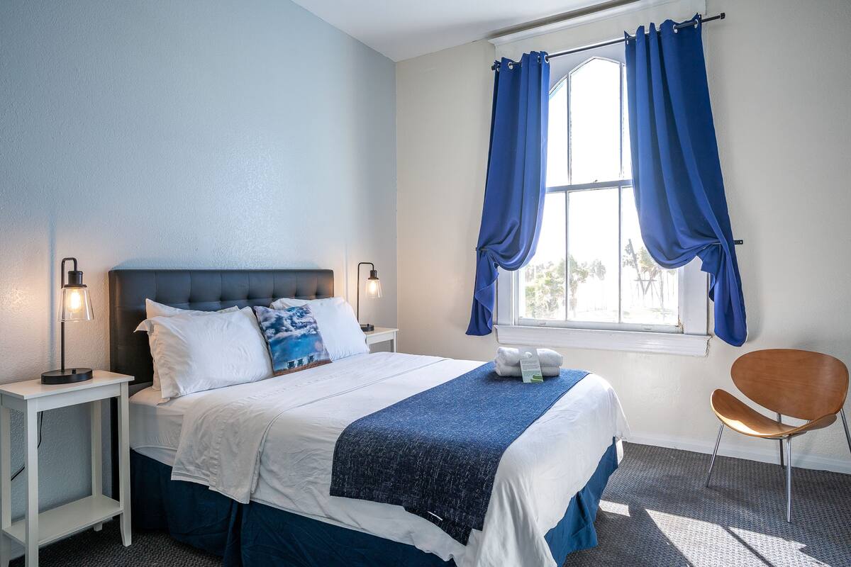 Un lit bien fait, recouvert d’une couverture bleue, une lampe de chevet et une chaise dans une pièce dotée d’une fenêtre et de rideaux.